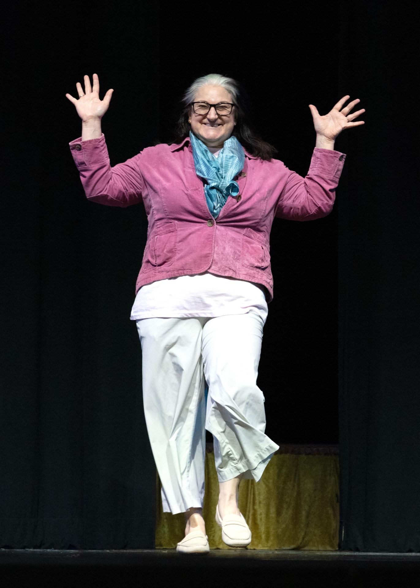 Marisa Laski smiling and waving on stage, wearing a pink jacket and blue scarf